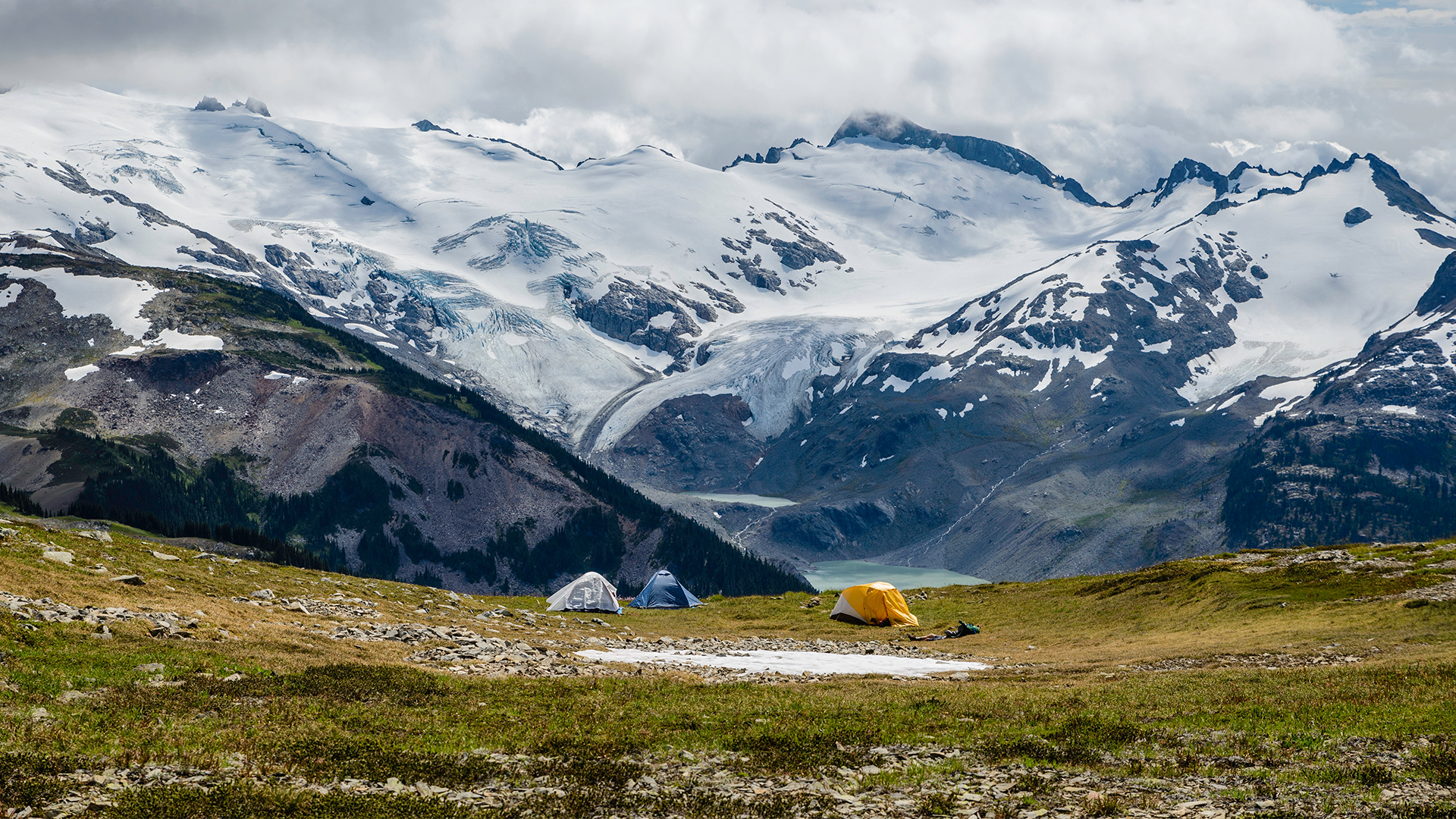 Snowy capped ridges in the background, three pitched tents on grass in the foreground